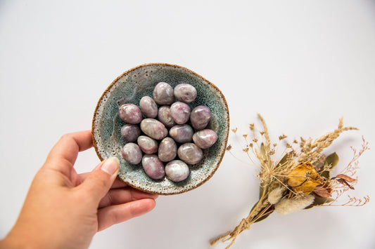 Hand holding a dish containing pink tourmlaine tumblestones with a posie of dried flowers in the background
